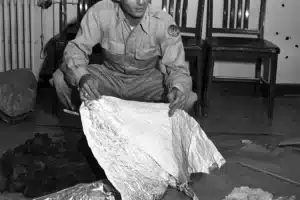 Man in a uniform crouches on the floor, handling a large crumpled foil-like sheet amid scattered debris in a room with chairs in the background.
