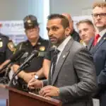 Man in a gray suit speaks at a podium with microphones, flanked by police officers and the U.S. flag behind him.