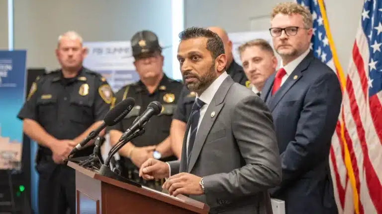 Man in a gray suit speaks at a podium with microphones, flanked by police officers and the U.S. flag behind him.