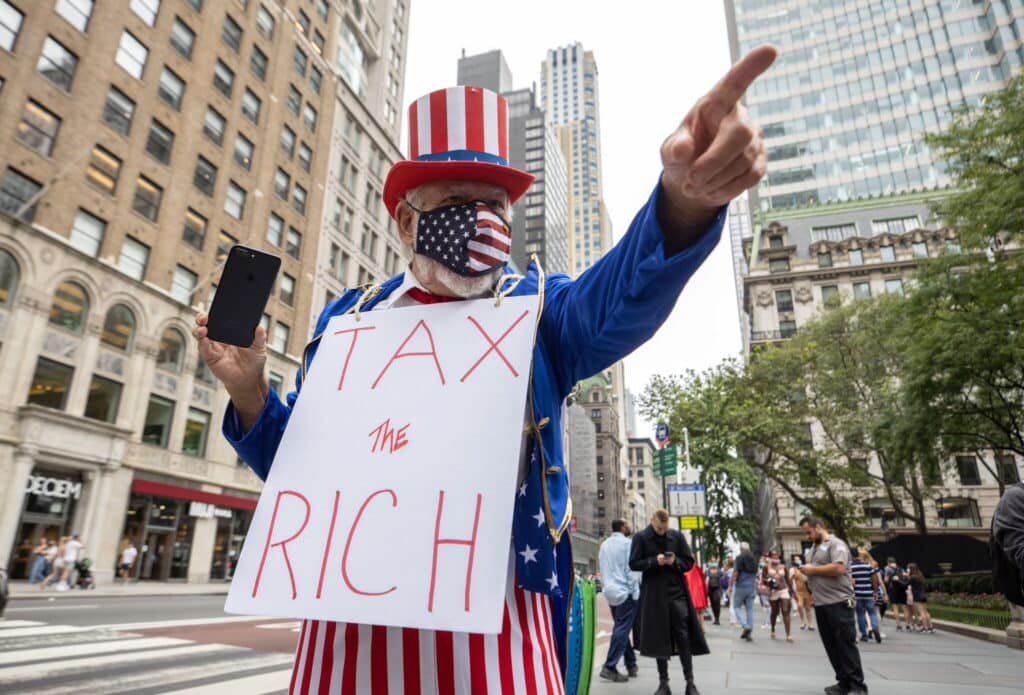 Protester dressed as Uncle Sam in a red‑white‑blue hat and star‑pattern mask holds a sign reading 'TAX THE RICH' on a city street.