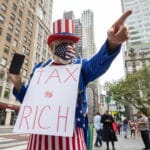 Protester dressed as Uncle Sam in a red‑white‑blue hat and star‑pattern mask holds a sign reading 'TAX THE RICH' on a city street.
