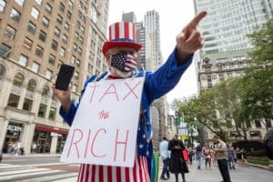 Protester dressed as Uncle Sam in a red‑white‑blue hat and star‑pattern mask holds a sign reading 'TAX THE RICH' on a city street.
