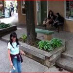 A woman in a light blue shirt and jeans walks along a sidewalk while two people sit on steps outside a storefront in the background.