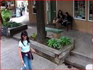 A woman in a light blue shirt and jeans walks along a sidewalk while two people sit on steps outside a storefront in the background.