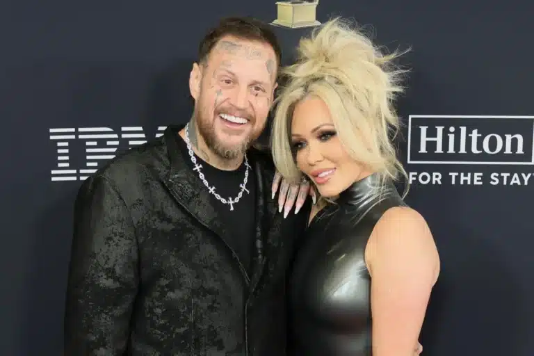 Smiling man with tattoos and a silver necklace posing with a platinum-haired woman in a metallic dress at a red carpet event, backdrop shows Hilton logo.