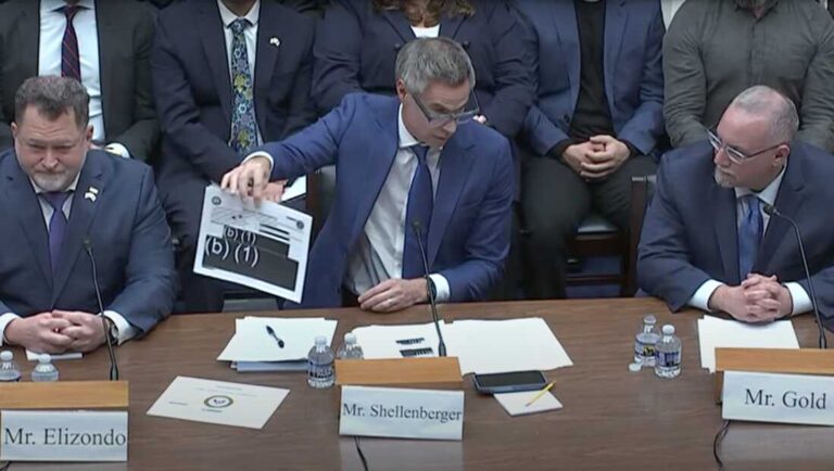Man in a blue suit presents a printed document at a formal meeting, with colleagues in suits nearby and microphones on the table.