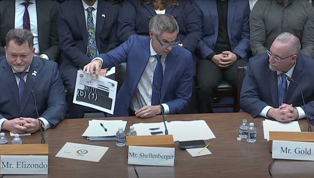 Man in a blue suit presents a printed document at a formal meeting, with colleagues in suits nearby and microphones on the table.