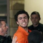 Man in an orange jumpsuit surrounded by several people indoors, some reaching toward him.