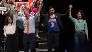 A diverse group on a stage raises their hands in victory, smiling, with campaign signs visible behind them.