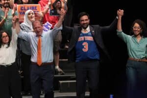 A diverse group on a stage raises their hands in victory, smiling, with campaign signs visible behind them.