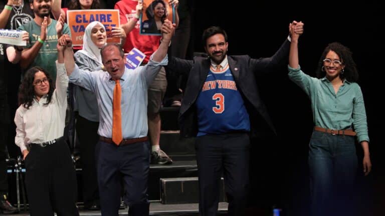 A diverse group on a stage raises their hands in victory, smiling, with campaign signs visible behind them.