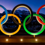Olympic rings sculpture with blue, yellow, black, green and red rings lit at dusk against mountains and a town backdrop.