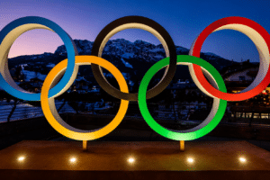 Olympic rings sculpture with blue, yellow, black, green and red rings lit at dusk against mountains and a town backdrop.