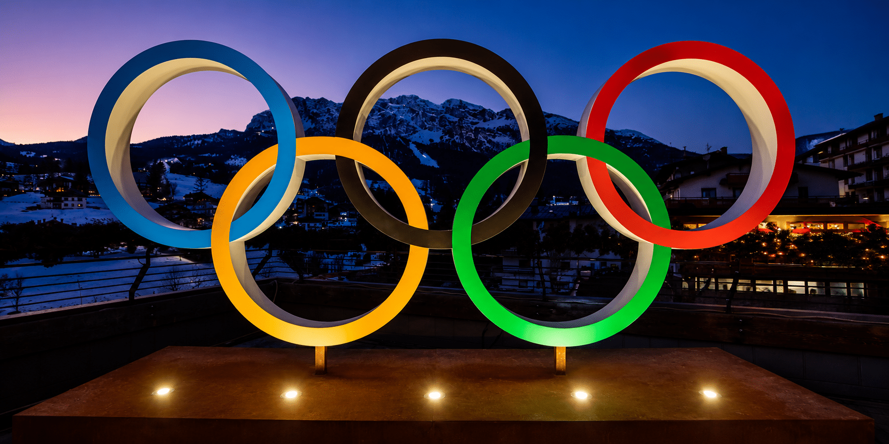 Olympic rings sculpture with blue, yellow, black, green and red rings lit at dusk against mountains and a town backdrop.
