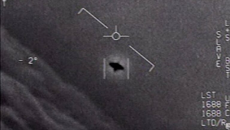 Grayscale medical image showing a circular crosshair marker with a diagonal line and a dark irregular shape near the lower center, plus measurement marks along the right edge.