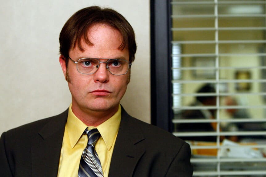 Stressed-looking man in a yellow shirt and suit stands in an office corridor with blinds behind him.