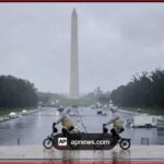 Two people ride small motorcycles on a wet, reflective pavement with the Washington Monument in the distance.
