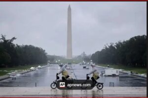 Two people ride small motorcycles on a wet, reflective pavement with the Washington Monument in the distance.