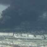 Industrial area with warehouses and rail yards, a large black smoke plume rising from a distant fire over the city edge.