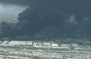 Industrial area with warehouses and rail yards, a large black smoke plume rising from a distant fire over the city edge.