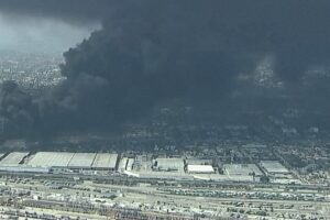 Industrial area with warehouses and rail yards, a large black smoke plume rising from a distant fire over the city edge.
