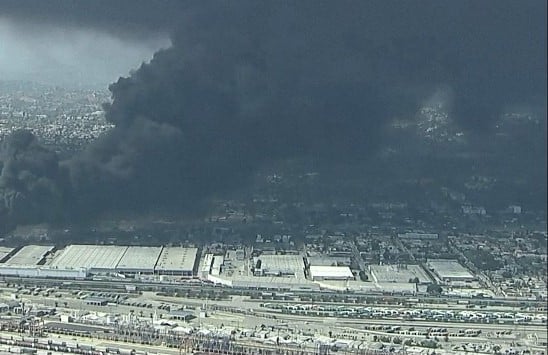 Industrial area with warehouses and rail yards, a large black smoke plume rising from a distant fire over the city edge.