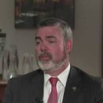 Middle-aged man with gray beard in a dark suit and red tie, speaking in an interview setting with wood paneling in the background.