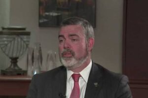 Middle-aged man with gray beard in a dark suit and red tie, speaking in an interview setting with wood paneling in the background.