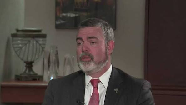 Middle-aged man with gray beard in a dark suit and red tie, speaking in an interview setting with wood paneling in the background.