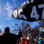 Person holds up a large black flag with white digits '9647' at a daytime protest; crowd and signs visible in the background under a sunny sky.