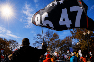 Person holds up a large black flag with white digits '9647' at a daytime protest; crowd and signs visible in the background under a sunny sky.
