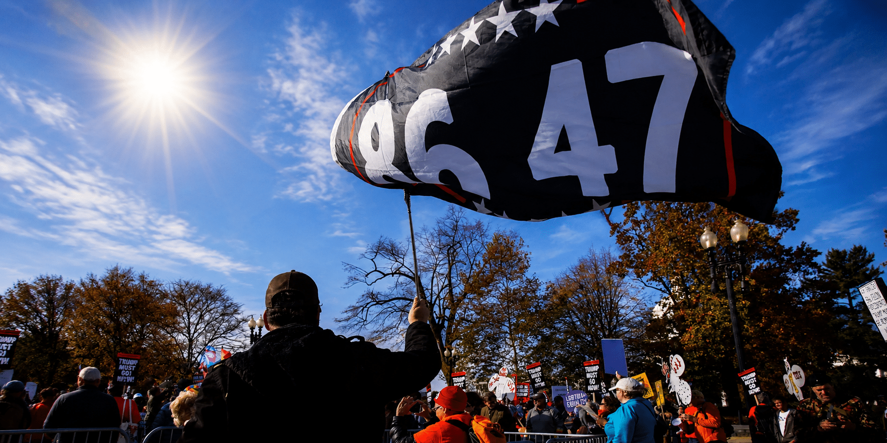 Person holds up a large black flag with white digits '9647' at a daytime protest; crowd and signs visible in the background under a sunny sky.