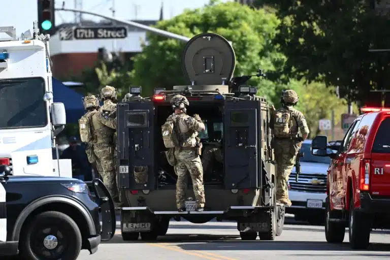 FBI tactical team in camouflage gear standing around a military-style armored vehicle on a city street, with police cars nearby.