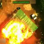 Aerial view of a large fireball explosion on the ground, with a green tarp-covered object nearby and trees along the edge; UNCLASSIFIED banner at the top.