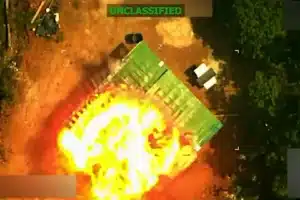 Aerial view of a large fireball explosion on the ground, with a green tarp-covered object nearby and trees along the edge; UNCLASSIFIED banner at the top.