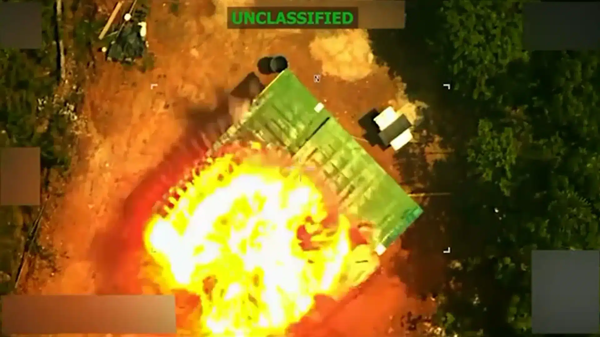 Aerial view of a large fireball explosion on the ground, with a green tarp-covered object nearby and trees along the edge; UNCLASSIFIED banner at the top.