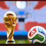Golden soccer trophy beside a colorful football on a green pitch with stadium seating in the background.