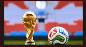 Golden soccer trophy beside a colorful football on a green pitch with stadium seating in the background.
