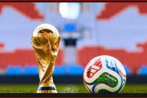 Golden soccer trophy beside a colorful football on a green pitch with stadium seating in the background.