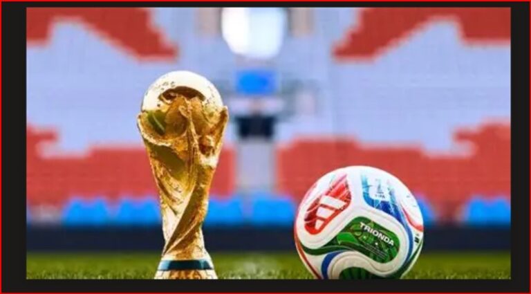 Golden soccer trophy beside a colorful football on a green pitch with stadium seating in the background.
