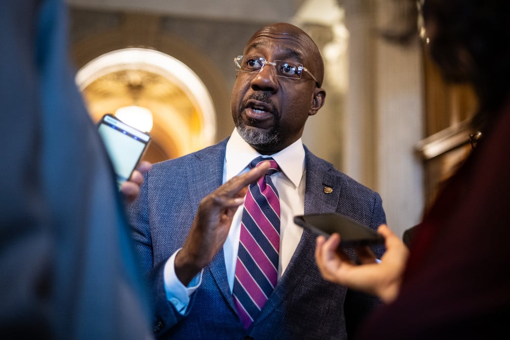 Man in a blue suit and pink striped tie speaks to reporters, surrounded by smartphones and recording devices.