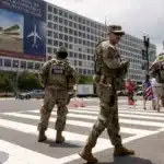 National Guard soldiers in camouflage uniforms stand in a crosswalk blocking traffic in front of a large office building with banners on the wall.