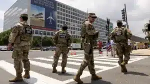 National Guard soldiers in camouflage uniforms stand in a crosswalk blocking traffic in front of a large office building with banners on the wall.