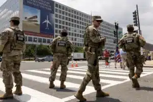 National Guard soldiers in camouflage uniforms stand in a crosswalk blocking traffic in front of a large office building with banners on the wall.