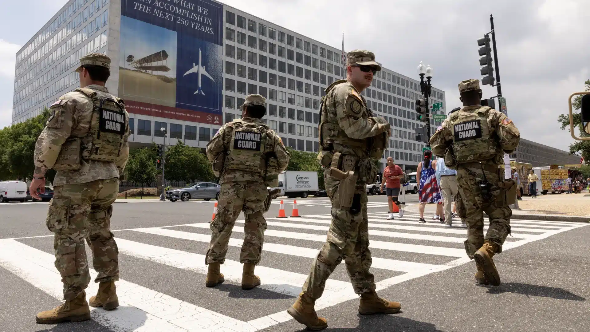 National Guard soldiers in camouflage uniforms stand in a crosswalk blocking traffic in front of a large office building with banners on the wall.
