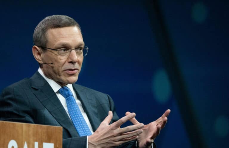 Man in a dark suit and blue tie speaks at a podium, wearing a headset microphone while gesturing with his hands on stage with a blue backdrop.