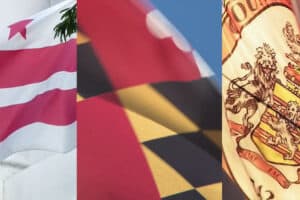 Three flags fluttering side by side in the wind: left has pink and white stripes, center shows a red flag with dark shapes, right displays a heraldic flag with a coat of arms.