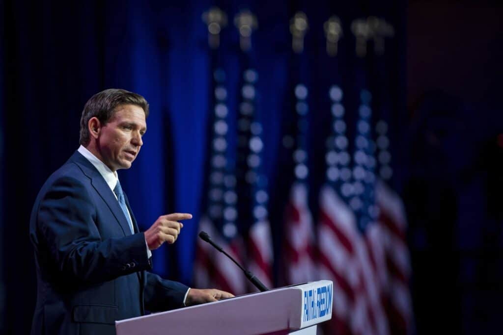 Man in a dark suit delivering a speech at a podium on a stage with American flags in the background.