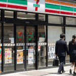 Two businesspeople walk past a 7-Eleven storefront with glass doors and Japanese signage outside the store intricate posters visible in the windows as pedestrians pass by.