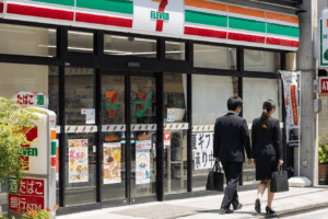 Two businesspeople walk past a 7-Eleven storefront with glass doors and Japanese signage outside the store intricate posters visible in the windows as pedestrians pass by.